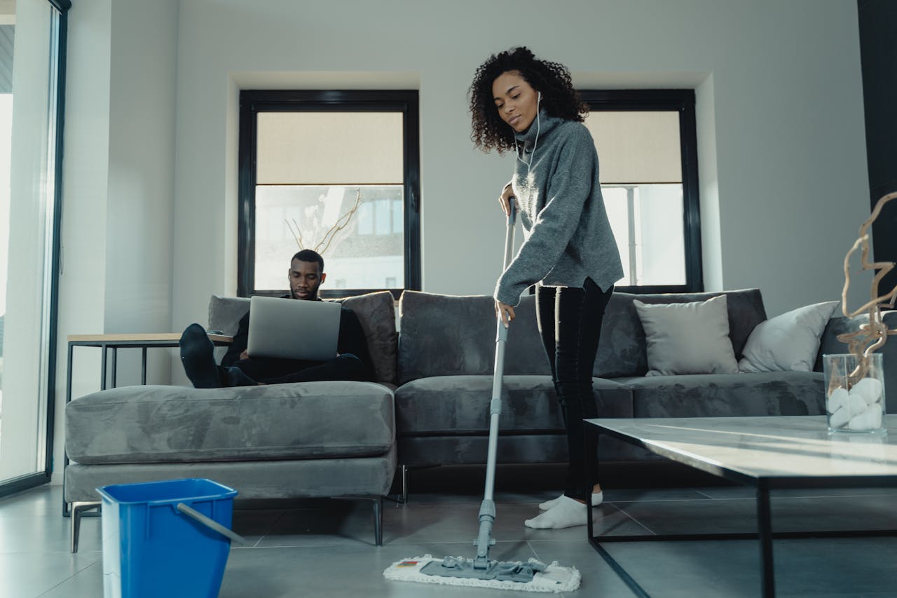 A woman mopping the floor while a man works on a laptop in a cozy modern living room.
